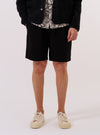 A model wears the Black Kelly Cotton Simple Short against a plain white background. The shorts feature a relaxed fit, an elasticated waistband, and side slant pockets. The black cotton fabric has a clean, smooth finish. The model is shown from the waist down, wearing the shorts with a white t-shirt and black shoes.