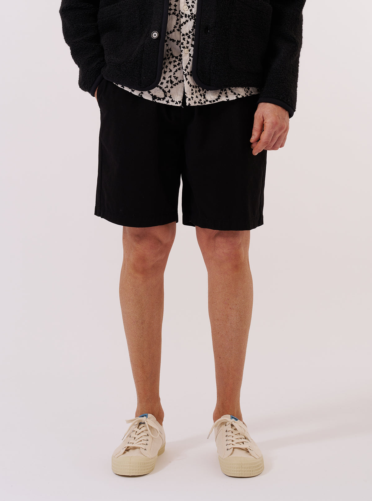 A model wears the Black Kelly Cotton Simple Short against a plain white background. The shorts feature a relaxed fit, an elasticated waistband, and side slant pockets. The black cotton fabric has a clean, smooth finish. The model is shown from the waist down, wearing the shorts with a white t-shirt and black shoes.