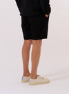 A model is wearing the Black Kelly Cotton Simple Short, shown from the waist down against a plain white background. The shorts feature a relaxed fit, an elasticated waistband, and side pockets. The black cotton fabric has a smooth, consistent finish. The model is wearing the shorts with a white t-shirt and white socks.