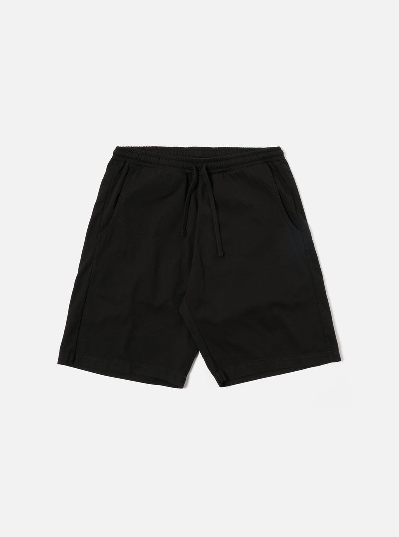 The Black Kelly Cotton Simple Short is shown as a flat lay against a white background. The shorts feature an elasticated waistband with a drawstring, two slanted side pockets, and a single rear patch pocket. The fabric is a solid black cotton with a slight texture.