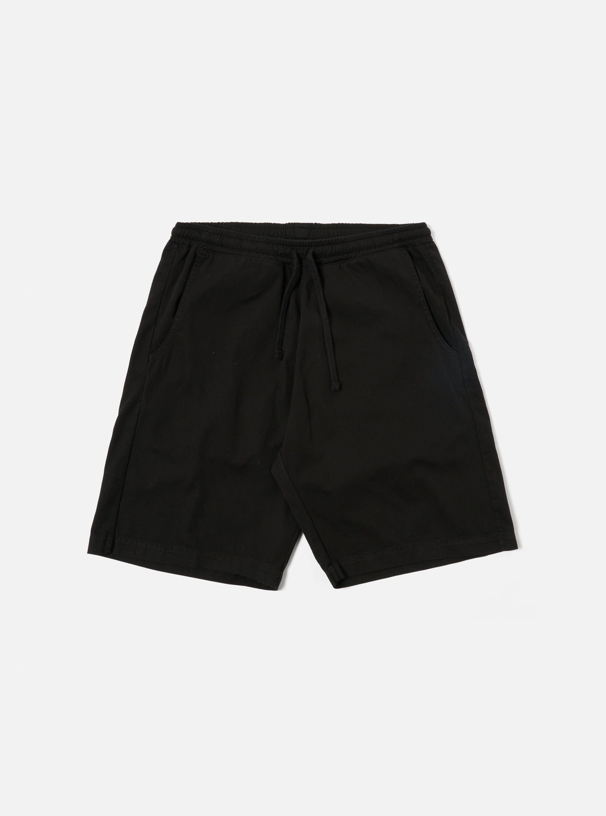 The Black Kelly Cotton Simple Short is shown as a flat lay against a white background. The shorts feature an elasticated waistband with a drawstring, two slanted side pockets, and a single rear patch pocket. The fabric is a solid black cotton with a slight texture.