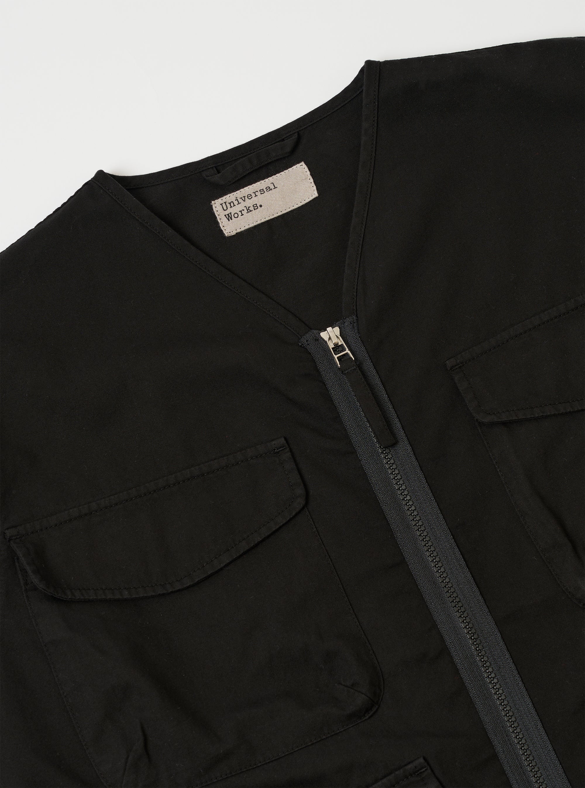 The Black Kelly Cotton Glider Jacket is shown from the back as it is worn by a model against a plain white background. The image highlights the clean, straight lines of the jacket's back panel and the bound edge detailing at the collar and hem. The model is also wearing olive green trousers.