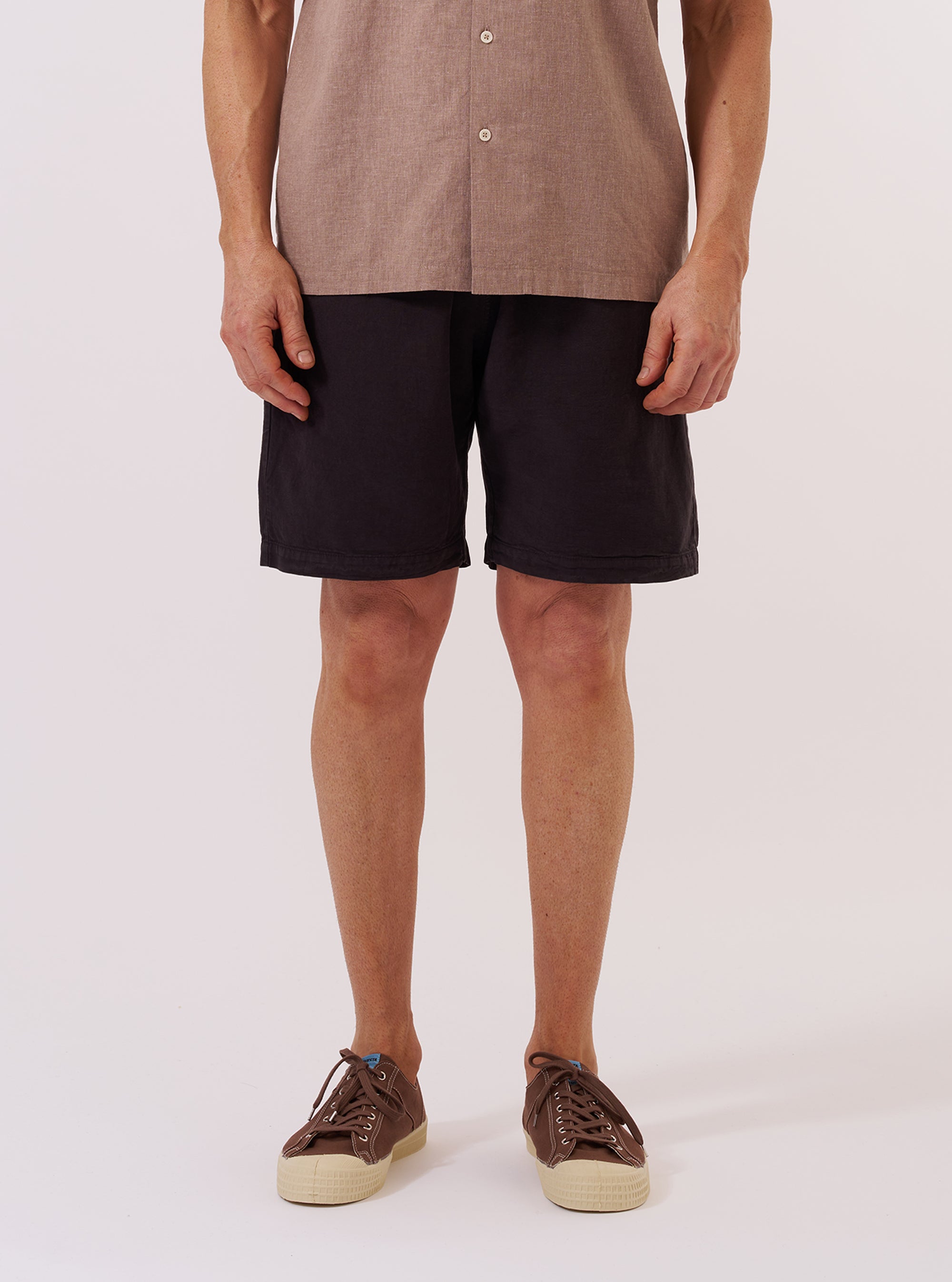 A model wears the Licorice Lyo Linen Mix Simple Short against a plain white background. The shorts feature a relaxed fit, an elasticated waistband with a drawstring, and side pockets. The fabric has a textured, lightweight appearance in a dark brown licorice color. The model is shown from the waist down, paired with a light-colored shirt and dark sandals.