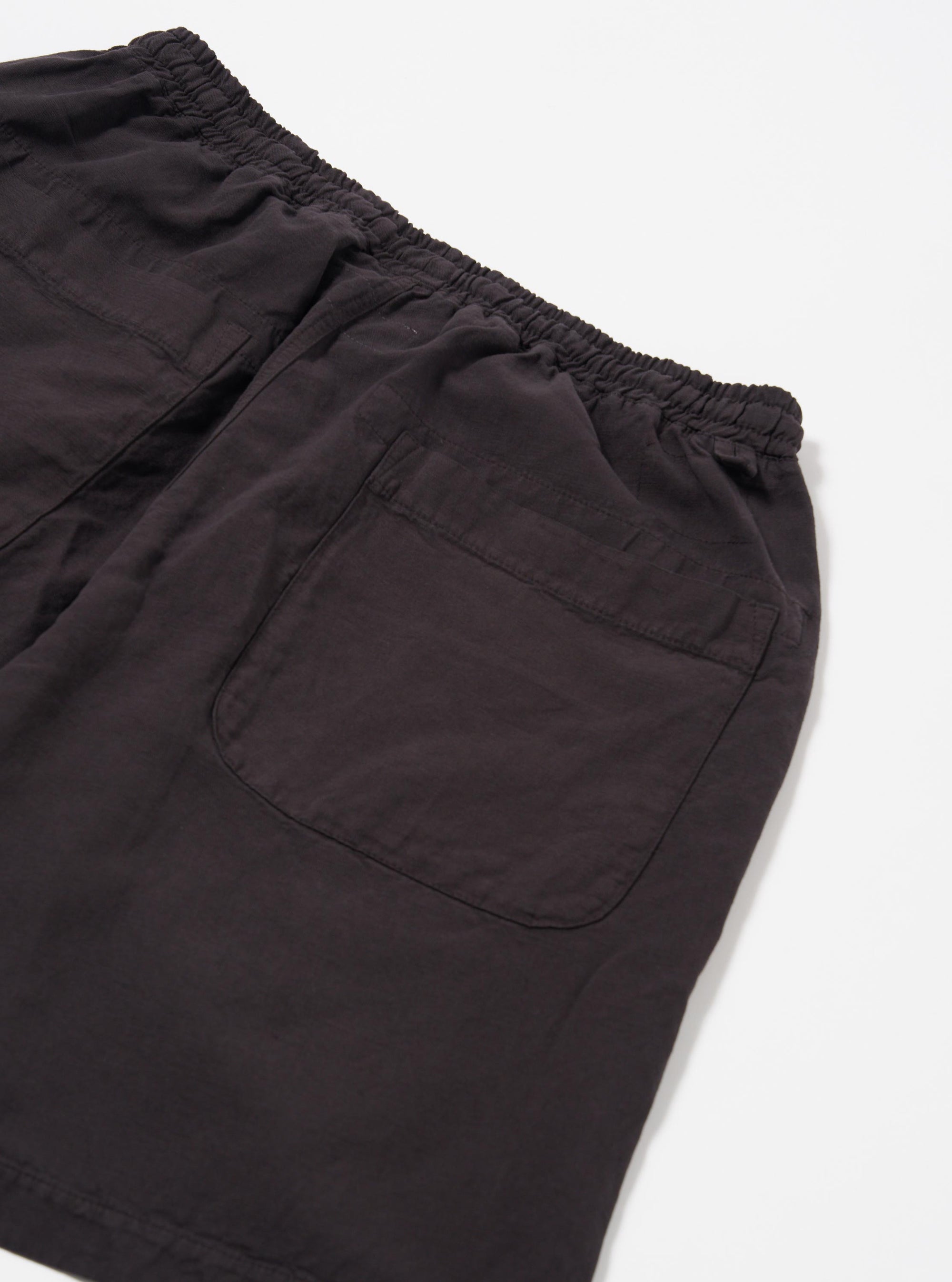 The Licorice Lyo Linen Mix Simple Short is displayed as a flat lay against a plain white background. The shorts feature an elasticated waistband with an internal drawstring, two side pockets, and a single rear buttoned flap pocket. The fabric has a textured appearance and is a dark licorice brown color.
