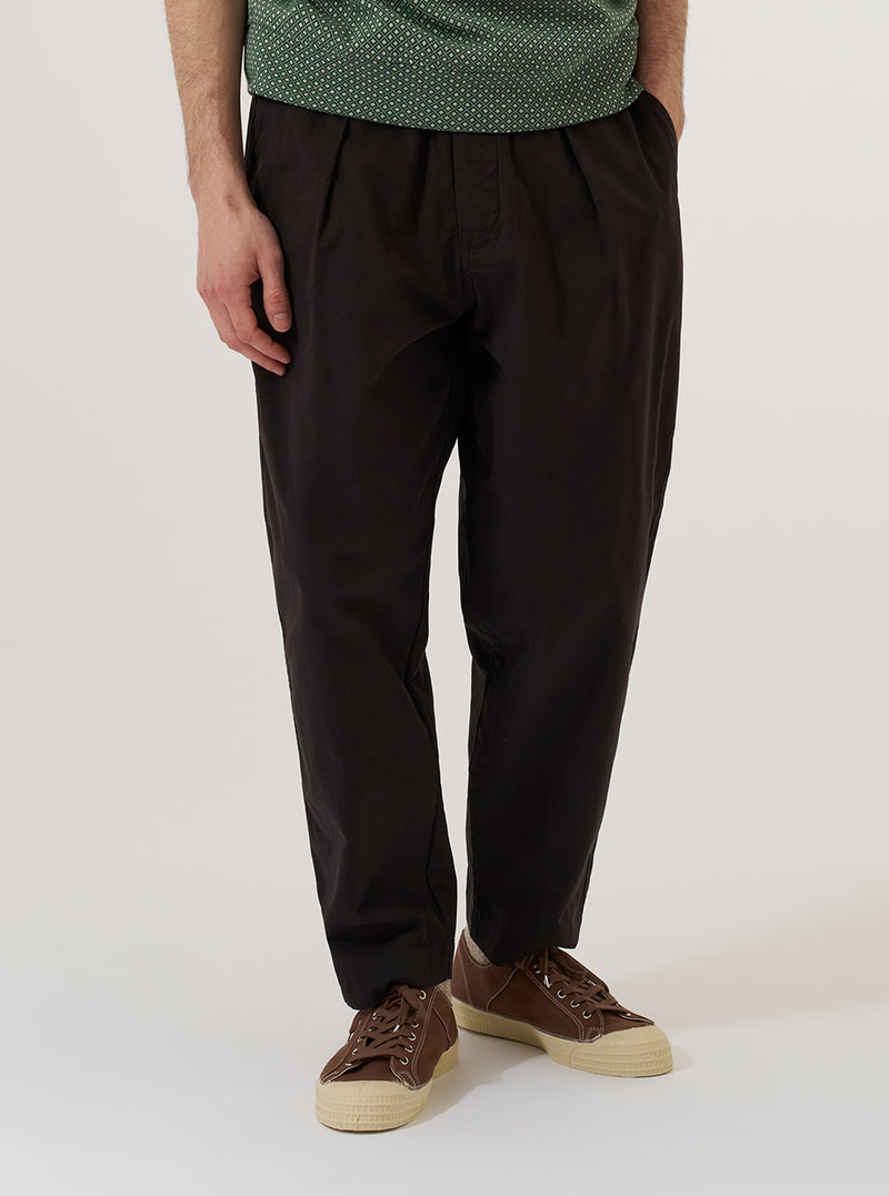 A model wears the Licorice Lyo Linen Mix Oxford Pant, shown from the waist down against a plain, neutral studio background. These loose-fit trousers in a deep brown licorice color feature a pleated front, a drawstring at the elasticated waistband, and side pockets. The model is wearing the pants with a tucked-in shirt and dark shoes, highlighting the relaxed, wide-leg silhouette.