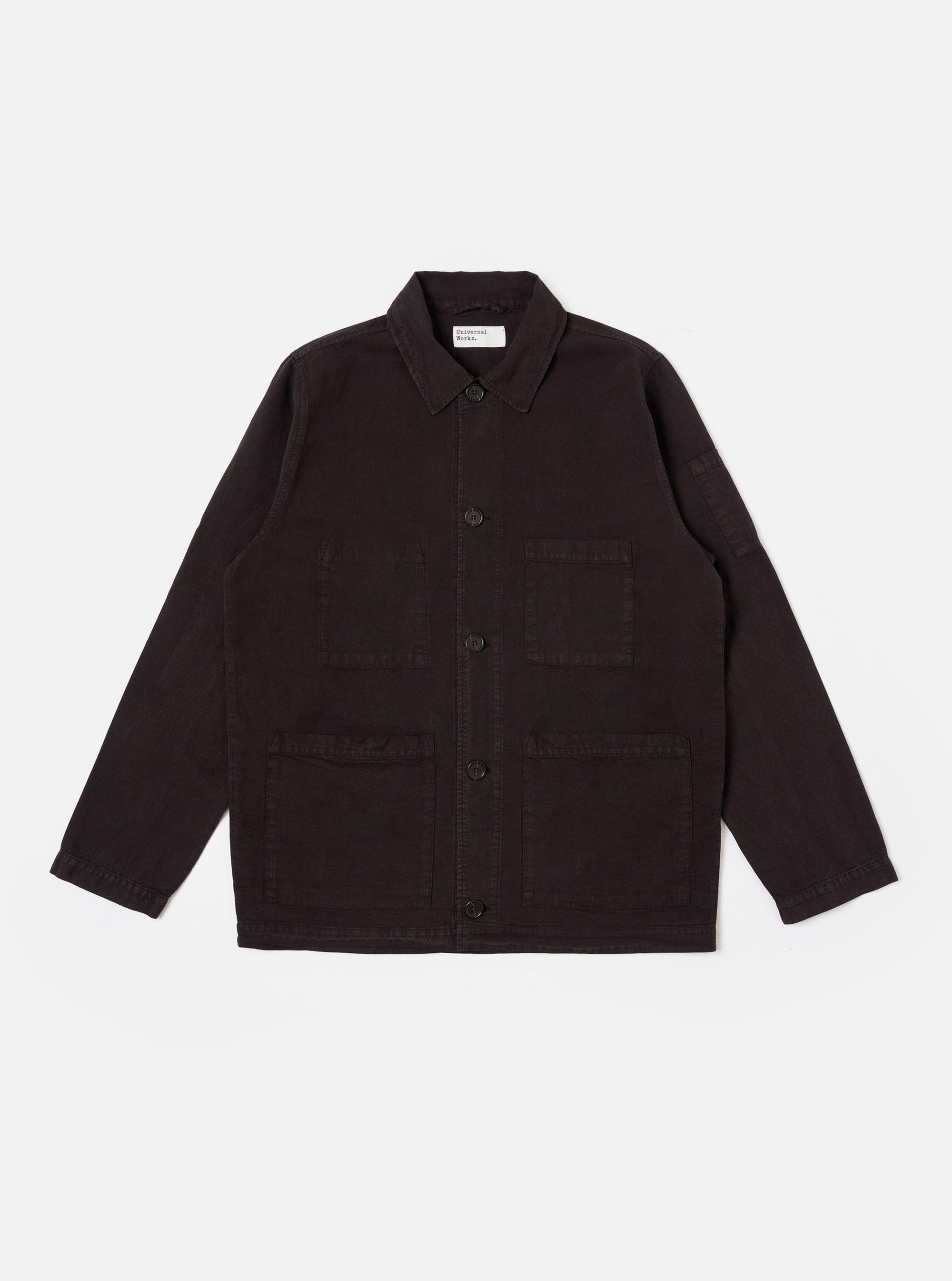 The Licorice Lyo Linen Mix Coverall Jacket is shown as a flat lay against a white background. The jacket features a full collar, a button-down front, and three large square patch pockets on the front, with an additional small pen pocket located on the sleeve. The garment is made from a brown lyo-linen mix fabric and has a short, square fit with simple cuffs.