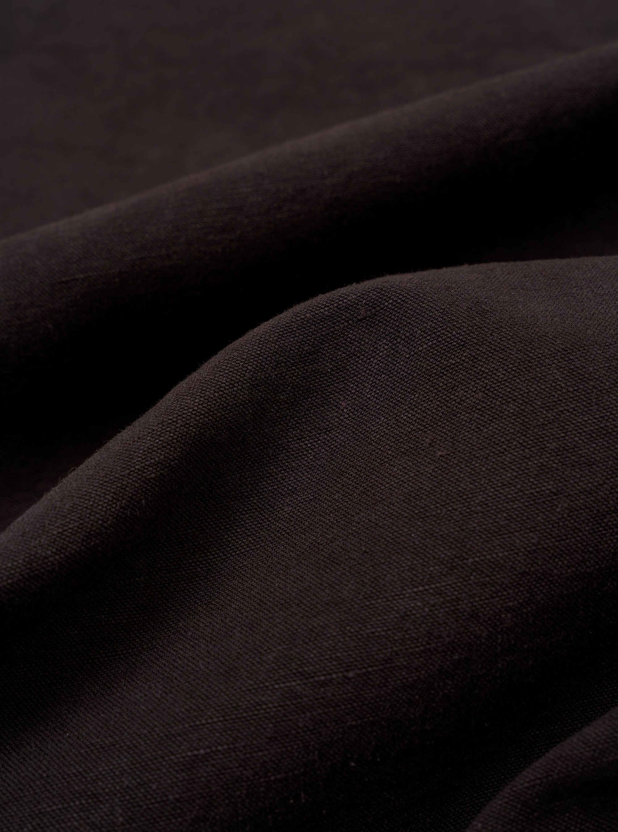 A close-up fabric swatch of the Licorice Lyo Linen Mix Coverall Jacket displays the dark brown color and the distinct texture of the linen, lyocell, and organic cotton blend. The material fills the frame, providing a detailed view of the fabric's weave.