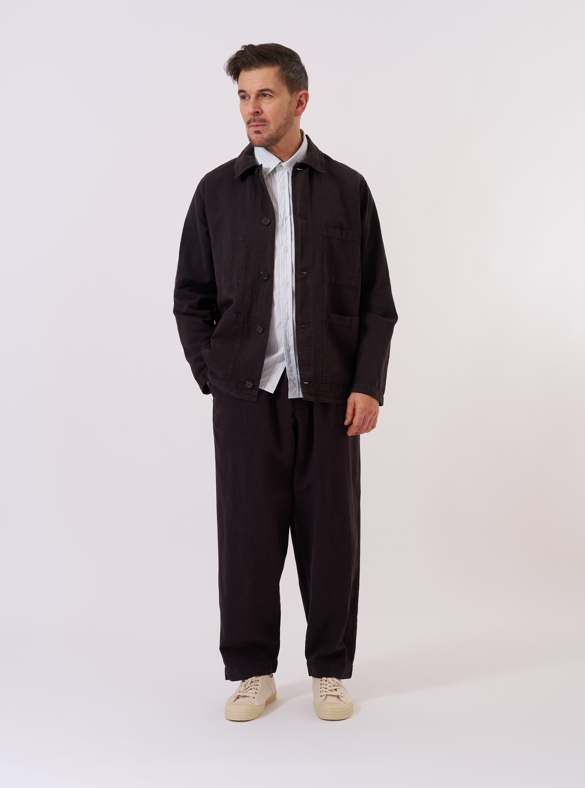 The Licorice Lyo Linen Mix Coverall Jacket is shown as a flat lay against a white background. This workwear-inspired jacket features a one-piece collar, a five-button front closure, and three square patch pockets on the front. A small pen pocket is visible on the upper left sleeve. The fabric is a smooth, solid dark brown blend.