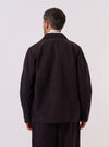 A model wears the Licorice Lyo Linen Mix Coverall Jacket, shown from the back against a plain white background. The jacket features a clean back panel with a central vertical seam, a straight hem, and a relaxed silhouette. The dark brown fabric highlights a smooth texture and a subtle drape characteristic of the linen and lyocell blend.