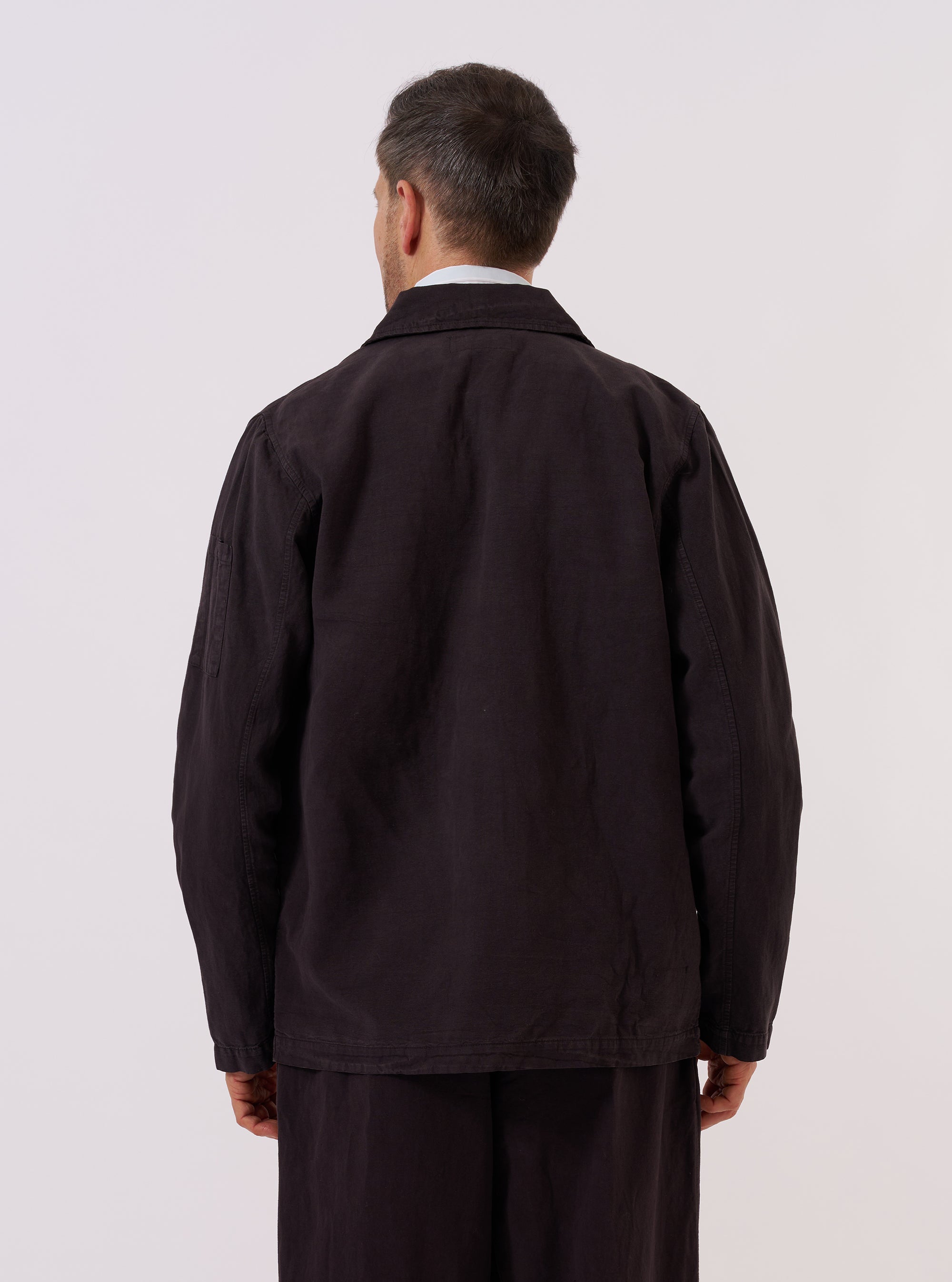 A model wears the Licorice Lyo Linen Mix Coverall Jacket, shown from the back against a plain white background. The jacket features a clean back panel with a central vertical seam, a straight hem, and a relaxed silhouette. The dark brown fabric highlights a smooth texture and a subtle drape characteristic of the linen and lyocell blend.
