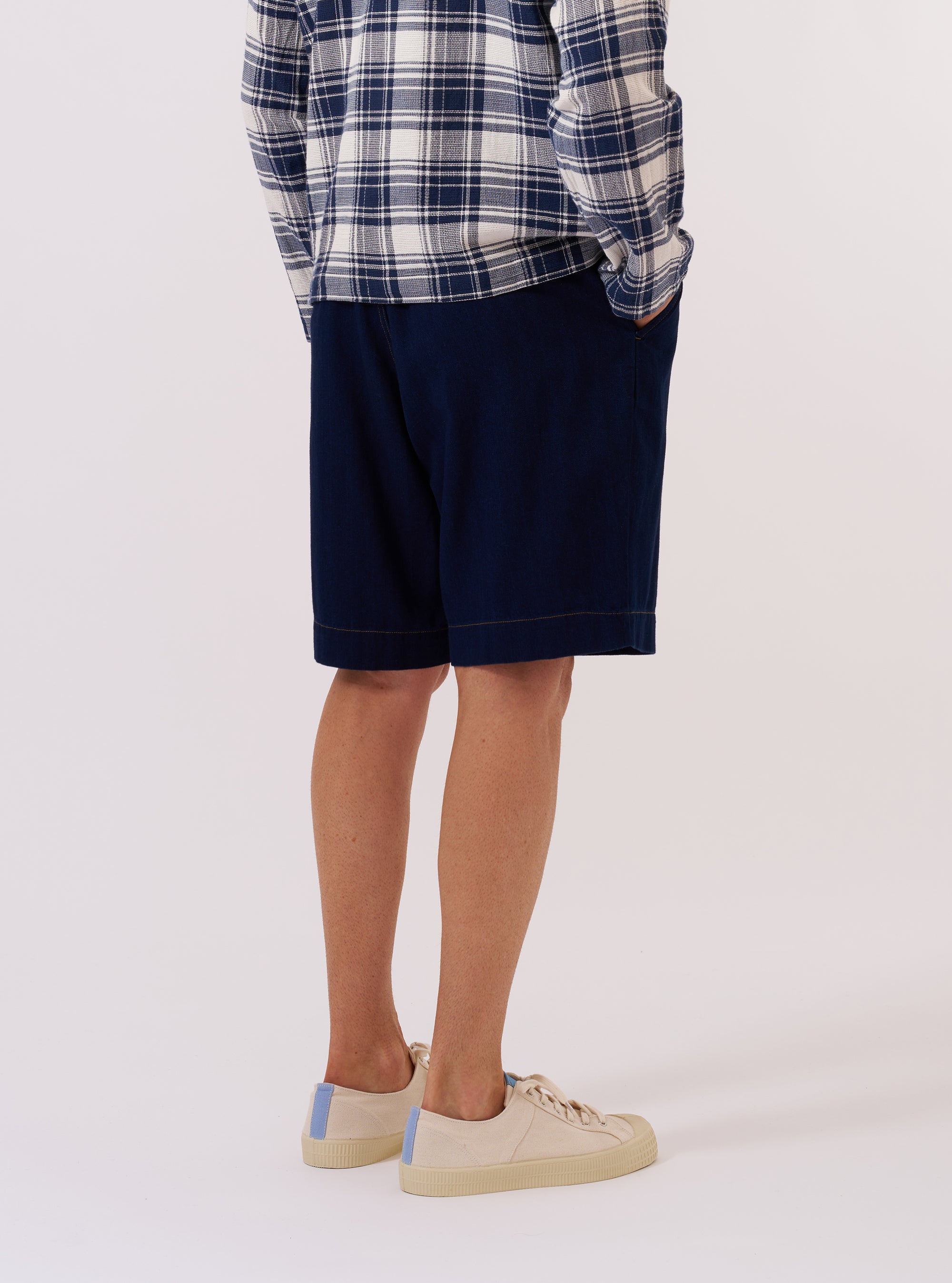A model wears the Indigo Recycled Herringbone Pleated Track Short against a plain, light-colored background. The shorts feature a visible herringbone texture, front pleats, and a relaxed fit. The model is shown from the waist down, showcasing the garment in a standing pose.