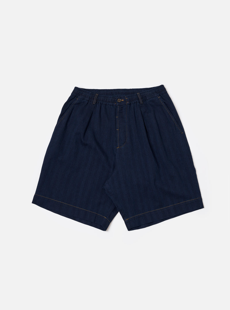 The Indigo Recycled Herringbone Pleated Track Short is shown as a flat lay against a white background. The shorts feature a visible herringbone weave pattern in indigo blue, front pleats, and an elasticated waistband with an adjustable drawstring.