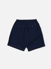 A model wears the Indigo Recycled Herringbone Pleated Track Short, shown from the waist down against a plain white background. The shorts feature a relaxed fit with a single pleat at the front and side pockets. The indigo-dyed fabric has a visible herringbone texture.