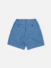The Washed Indigo Recycled Herringbone Pleated Track Short is shown from the back as a flat lay against a plain white background. The image highlights the rear construction, featuring one buttoned flap pocket on the left and one buttoned jetted pocket on the right. The elasticated waistband and the textured herringbone weave of the washed indigo fabric are clearly visible.