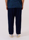 A model wears the Indigo Recycled Herringbone Pleated Track Pant against a plain white background. The image shows the model from the waist down, highlighting the loose, tapered fit and the single pleat at the front of each leg. The dark indigo fabric features a distinct herringbone weave. The model is wearing the trousers with white socks and dark footwear.