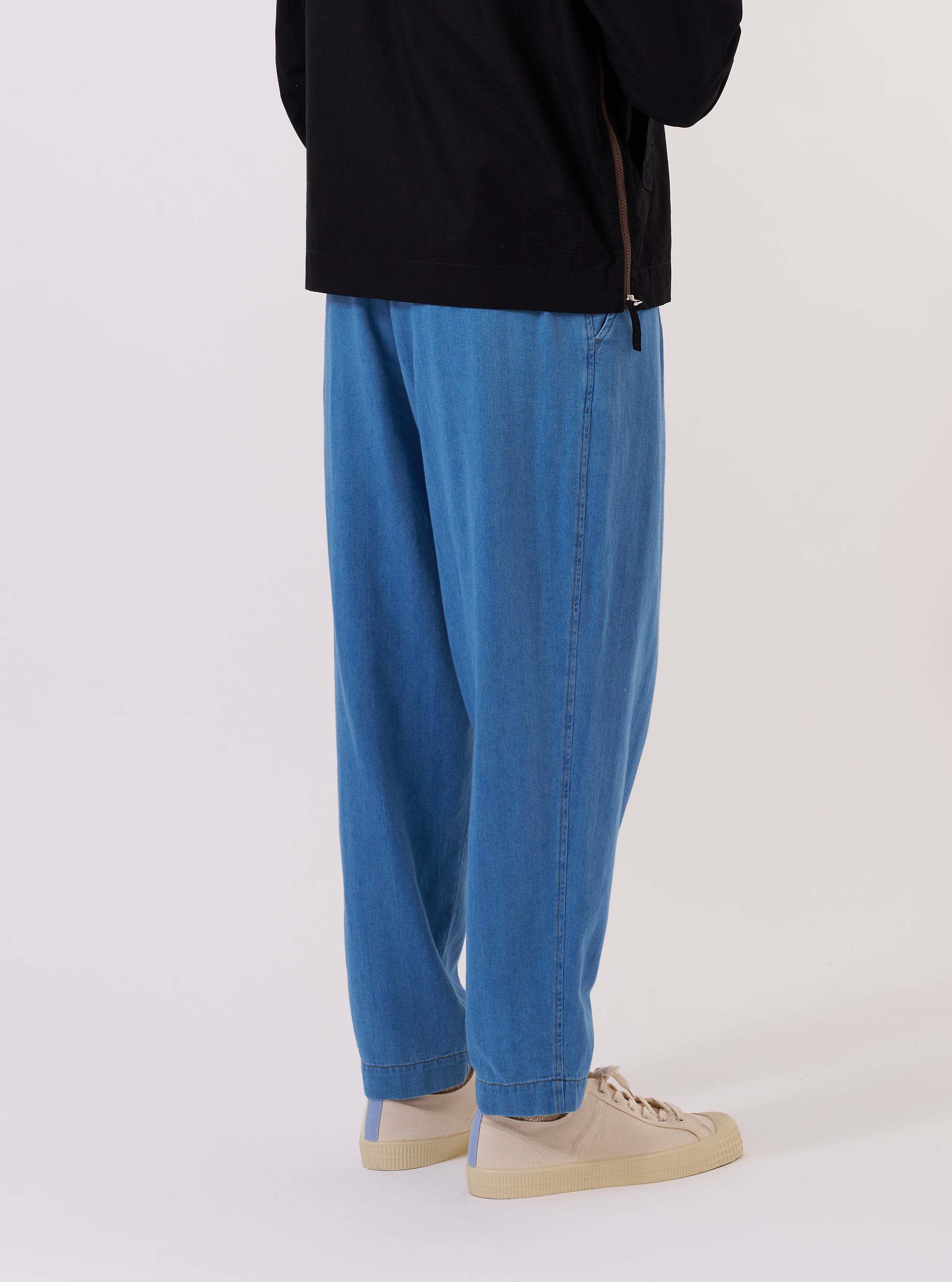 A model wears the Washed Indigo Recycled Herringbone Pleated Track Pant, shown from the rear three-quarter angle against a plain white background. The image highlights the loose, tapered fit and the textured herringbone weave of the washed indigo fabric. One rear patch pocket is visible on the right side, and the elasticated waistband is prominent. The model is also wearing a light-colored long-sleeve shirt and dark shoes.