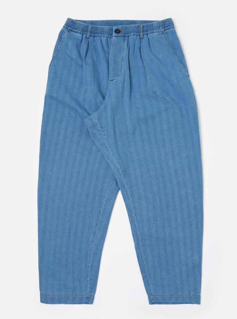 The Washed Indigo Recycled Herringbone Pleated Track Pant is shown as a flat lay against a white background. The trousers feature an elasticated waistband with an external drawstring, a single pleat at the front, and a loose, tapered leg. Two side pockets are visible, and the fabric displays a distinct herringbone texture in a washed indigo blue.