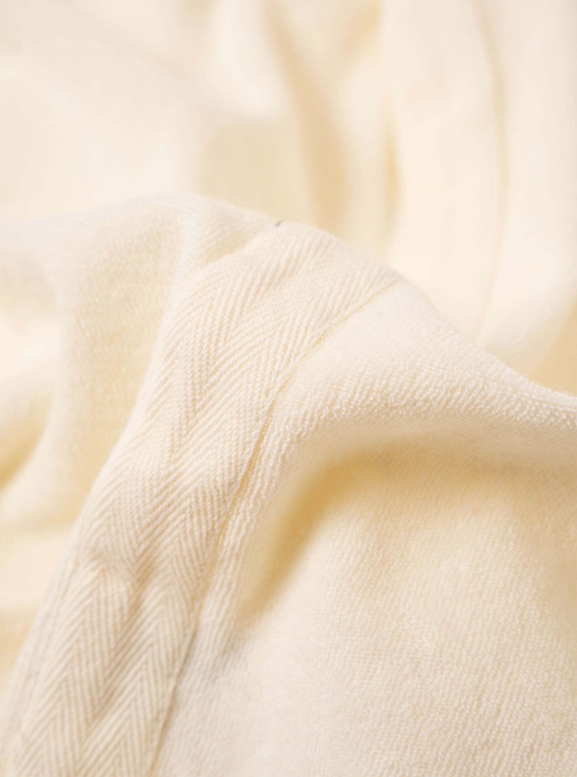 The Ecru Light Weight Terry Newlyn Polo is shown in a close-up detail shot against a plain white background. The image focuses on the soft, looped texture of the ecru terry cloth fabric, highlighting the clean stitching of the hem and the side of the garment.