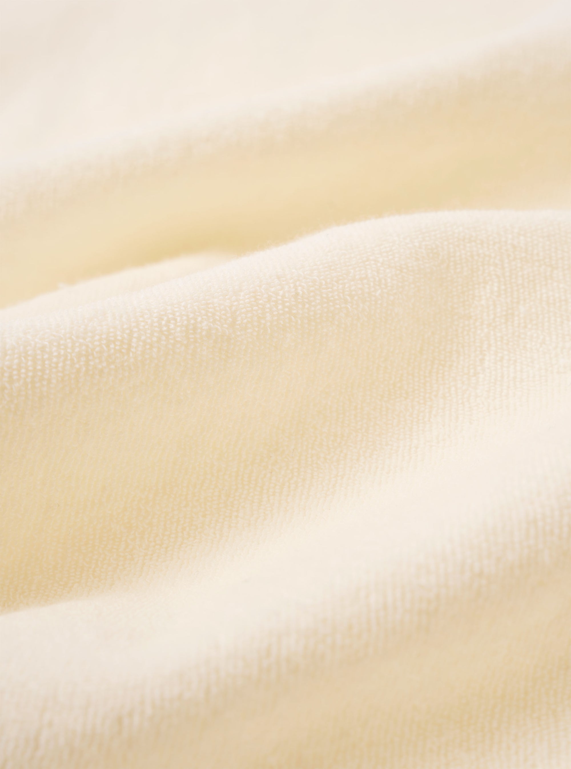 A close-up fabric swatch of the Ecru Light Weight Terry Newlyn Polo highlights the off-white color and the soft, looped texture of the lightweight terry cloth. The material fills the frame, providing a detailed view of the garment's pile and construction.