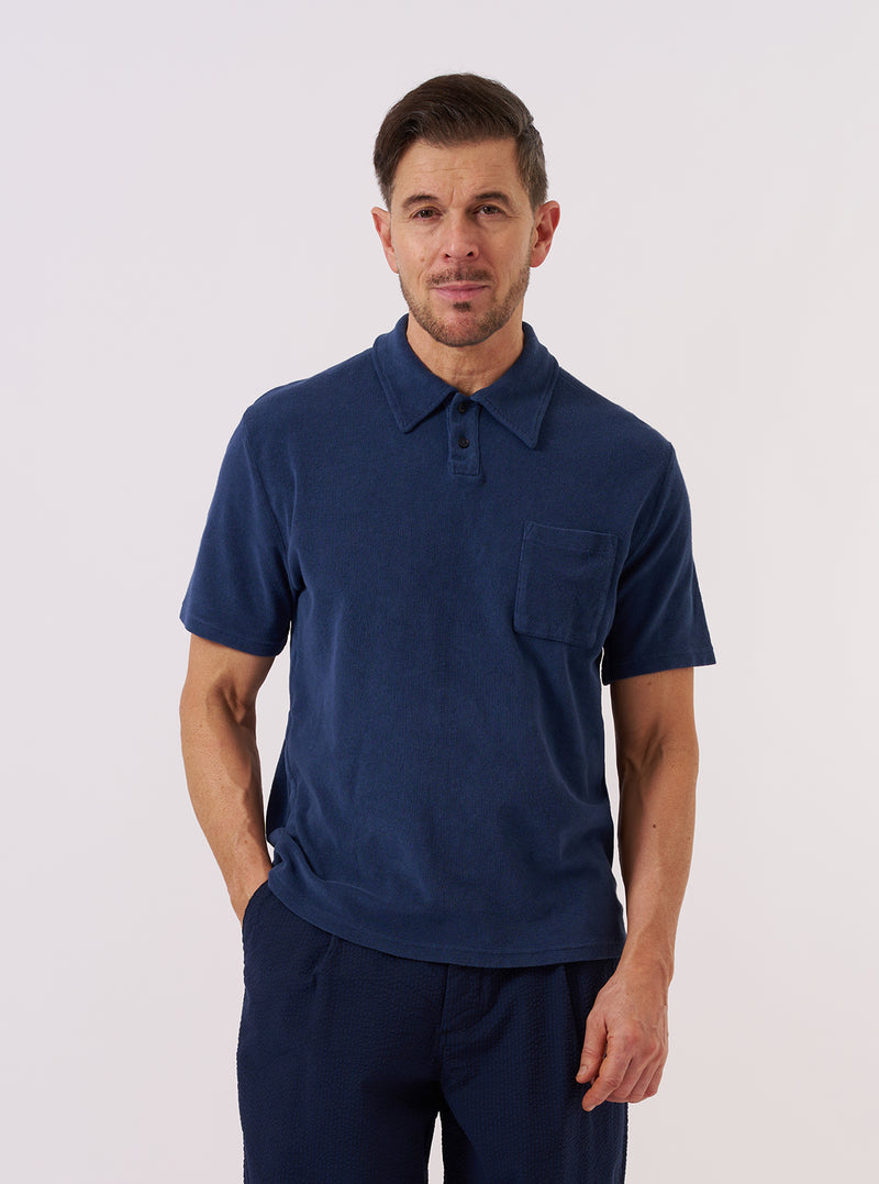 A model wears the Navy Light Weight Terry Newlyn Polo against a plain white background. The polo features a relaxed fit, a self-fabric one-piece collar, a two-button placket, and a large patch pocket on the left chest. The navy blue fabric has a soft, looped terry texture. The model is shown from the front, highlighting the garment's simple, overhead construction and split hem.