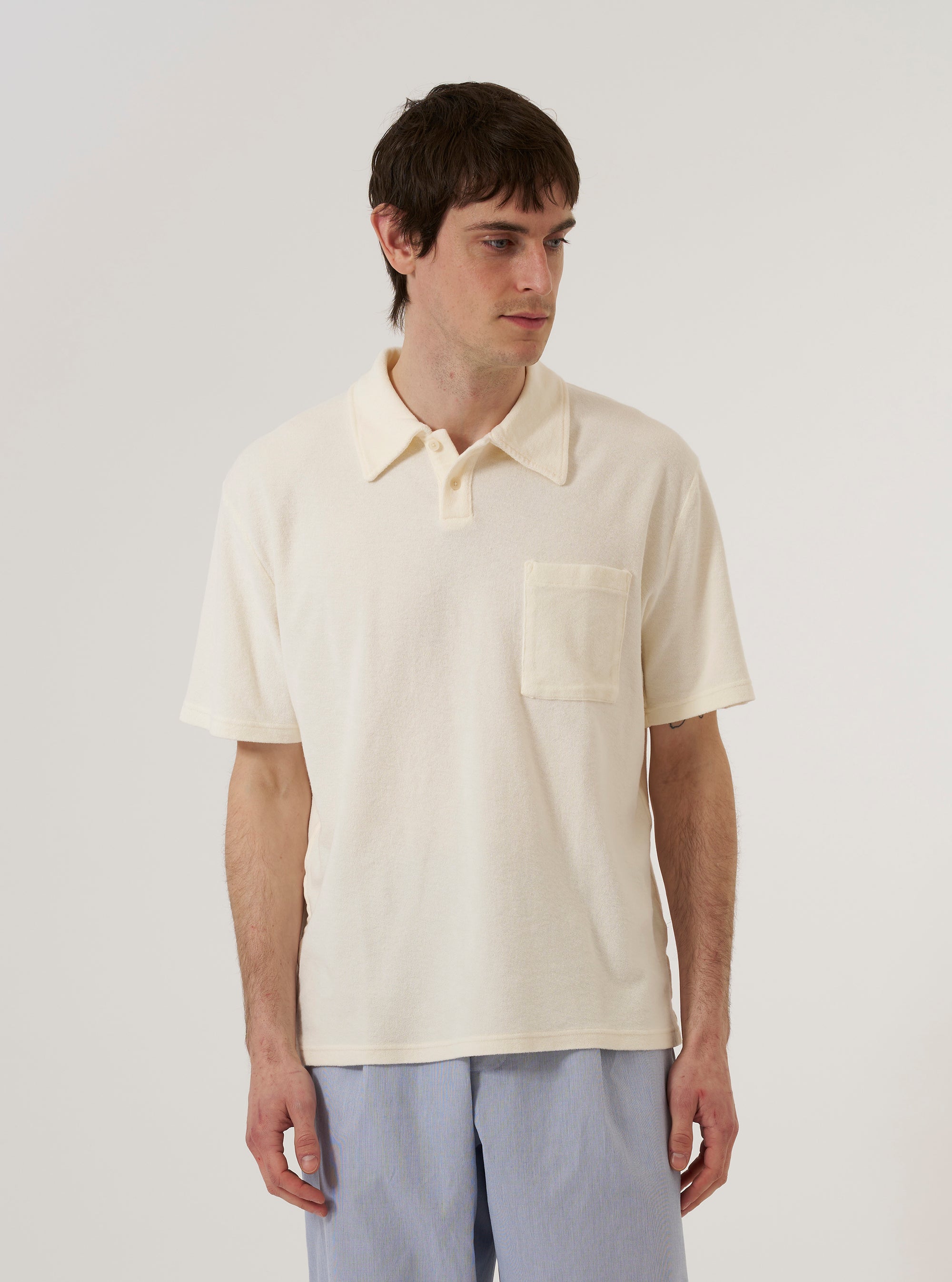 A model wears the Ecru Light Weight Terry Newlyn Polo against a plain white background. The polo features a three-button closure, a classic polo collar, and a single patch pocket on the chest. The ecru fabric has a soft, textured terry finish.