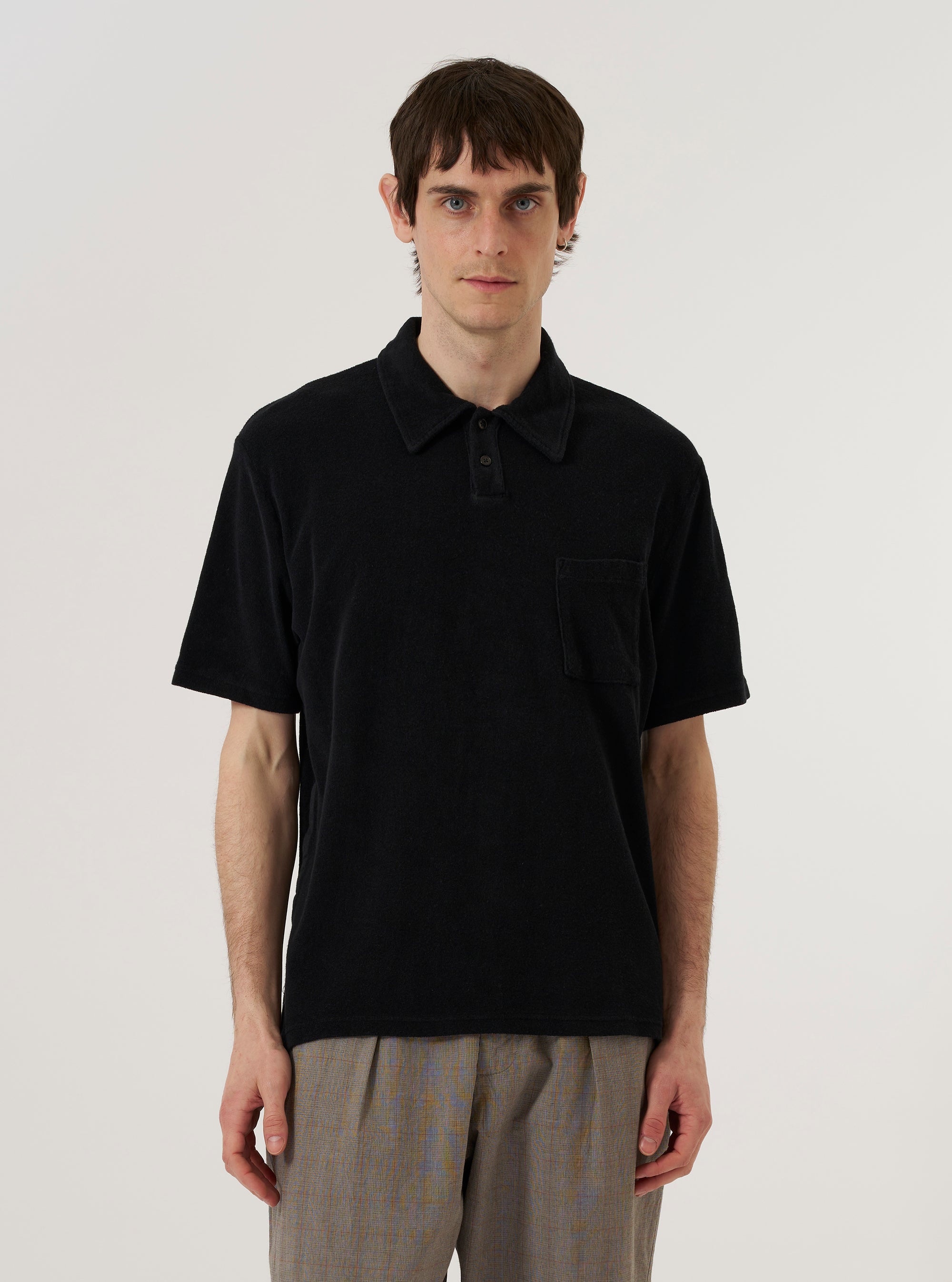 A model wears the Black Light Weight Terry Newlyn Polo against a plain white background. The polo shirt features a classic collar, a three-button placket, and short sleeves. The fabric is a solid black with a soft, textured light-weight terry finish. The model is shown from the waist up in a front-facing view.