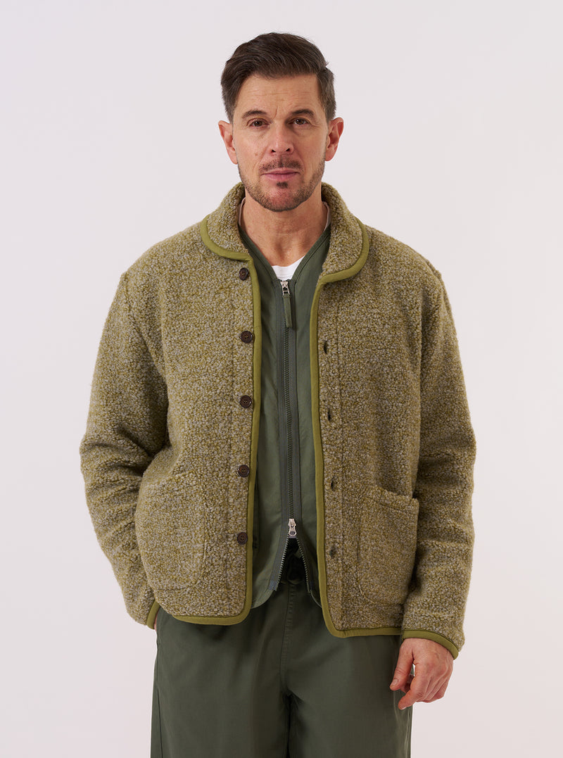 A model wears the Light Olive Alvar Fleece Lancaster Jacket against a plain white background. The jacket features a rounded shawl collar, a six-button front closure, and two large patch pockets at the waist. The light olive fabric has a soft, textured fleece appearance. The model is wearing the jacket layered over a light-colored long-sleeved shirt and dark trousers.