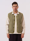 A model wears the Light Olive Alvar Fleece Lancaster Gilet over a light-colored long-sleeve shirt, standing against a plain, neutral background. The gilet features a textured, curly fleece fabric, a V-neckline, a central button-down closure, and two large patch pockets at the waist.