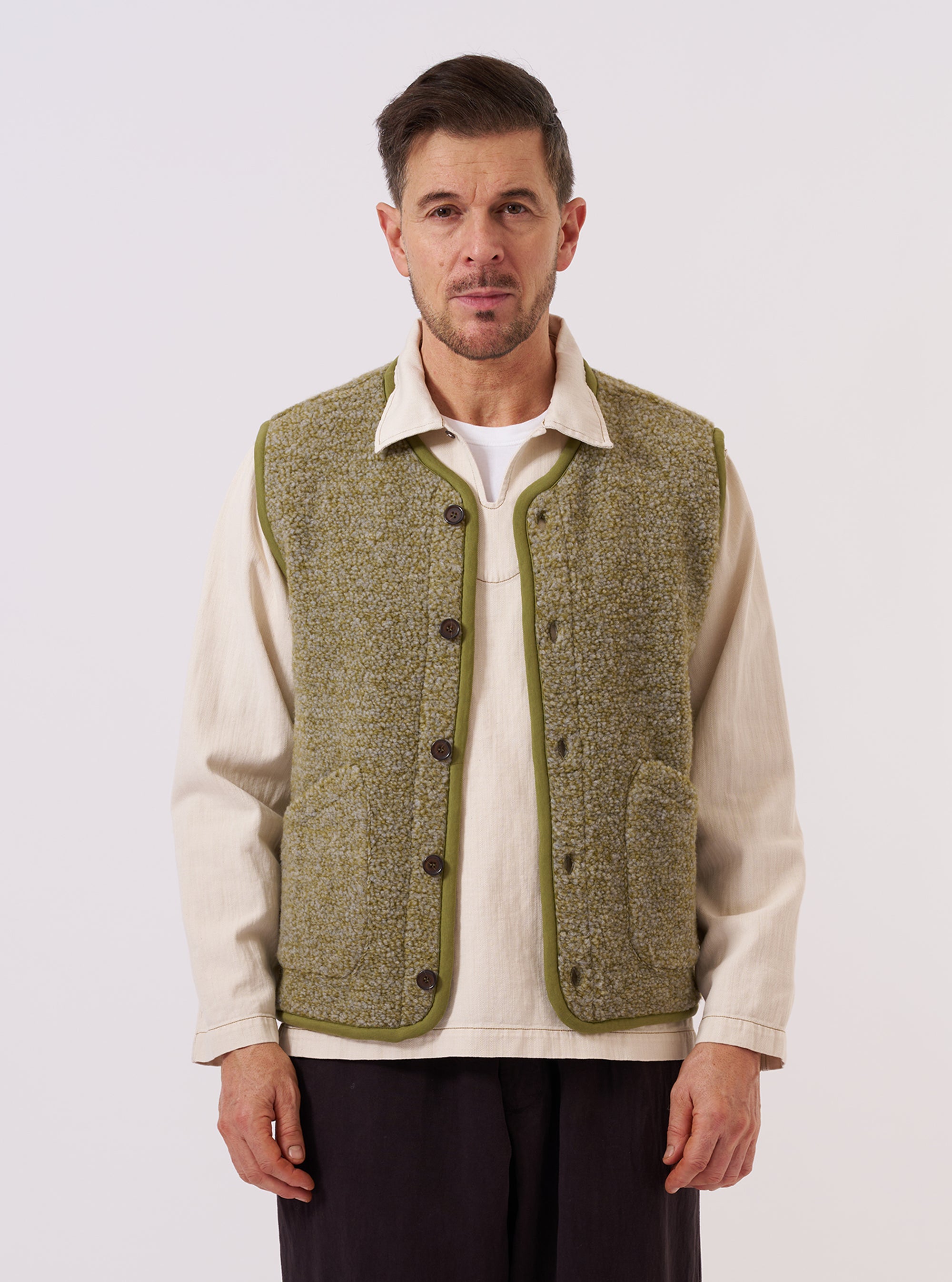 A model wears the Light Olive Alvar Fleece Lancaster Gilet over a light-colored long-sleeve shirt, standing against a plain, neutral background. The gilet features a textured, curly fleece fabric, a V-neckline, a central button-down closure, and two large patch pockets at the waist.