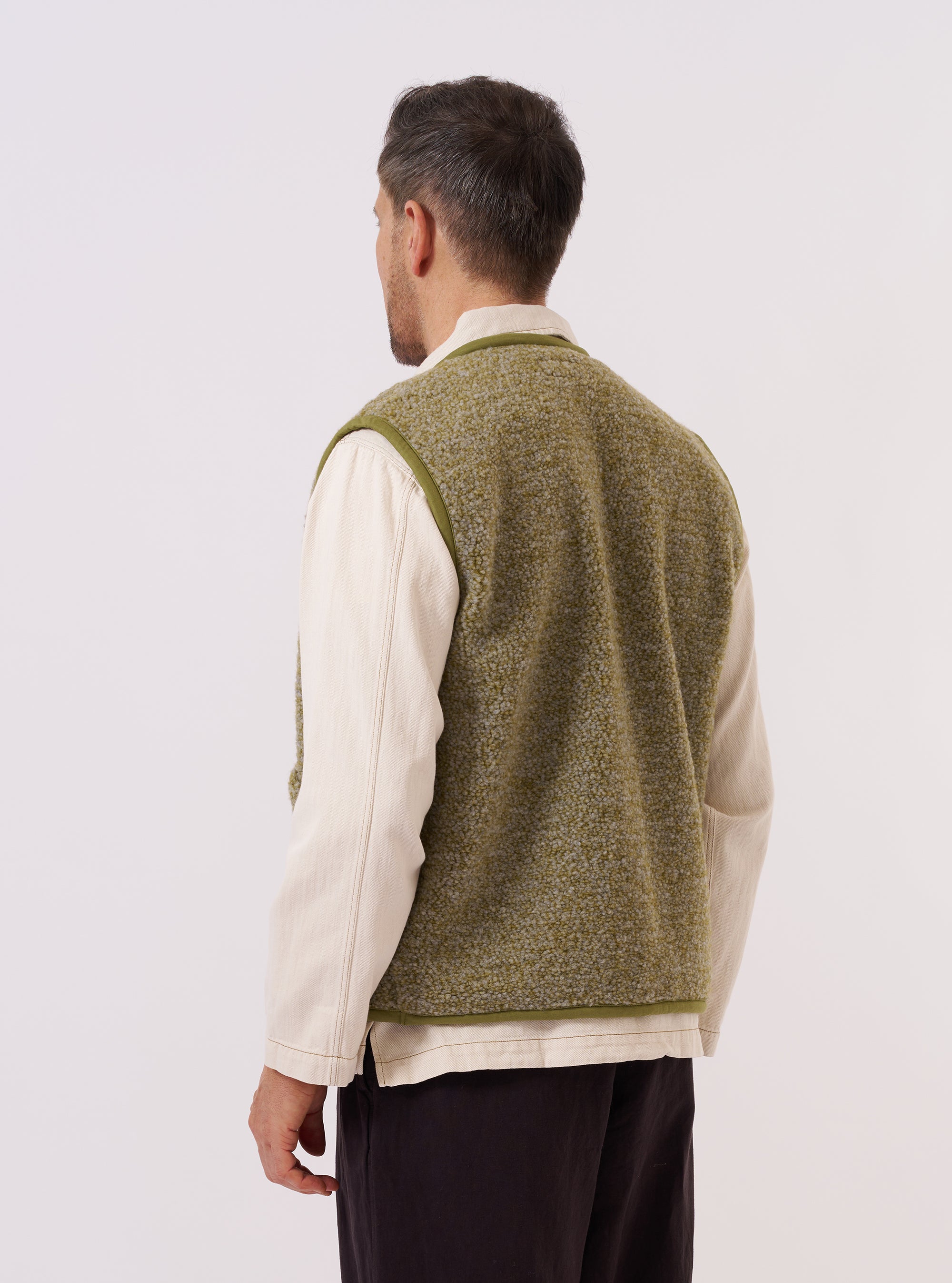 A model wears the Light Olive Alvar Fleece Lancaster Gilet over a light-colored shirt, shown from a rear three-quarter angle against a plain white background. The gilet features a sleeveless design with a straight hem and is made from a textured, light olive green fleece fabric. The model is standing with their back partially turned to the camera to highlight the garment's fit and rear construction.