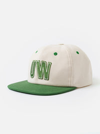 The Ecru/Green Canvas Baseball Hat is shown as a flat lay against a plain white background. The hat features a dual-tone design with an ecru crown and a contrasting green curved brim. A small, circular ventilation eyelet is visible on the side of the crown, and a matching green top button completes the design.
