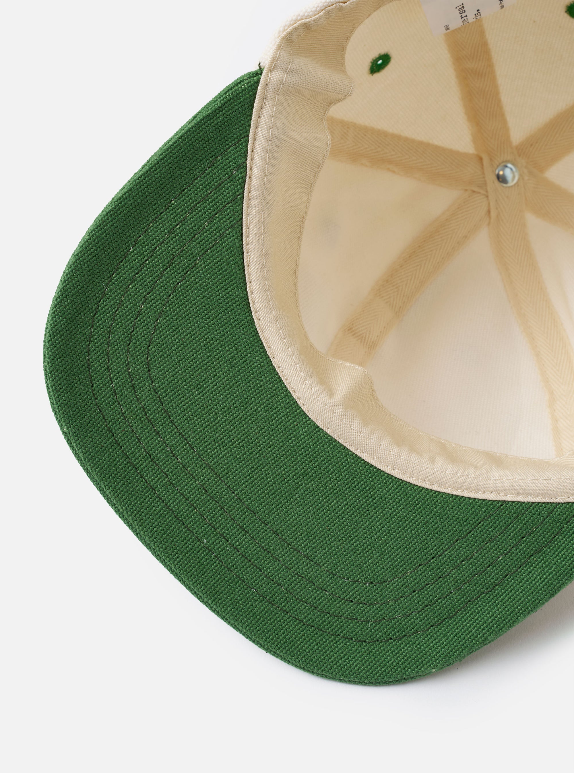 The Ecru/Green Canvas Baseball Hat is shown as a flat lay from the side against a plain white background. The hat features a structured six-panel crown in ecru canvas and a curved green brim. A metal eyelet is visible on the side panel, and