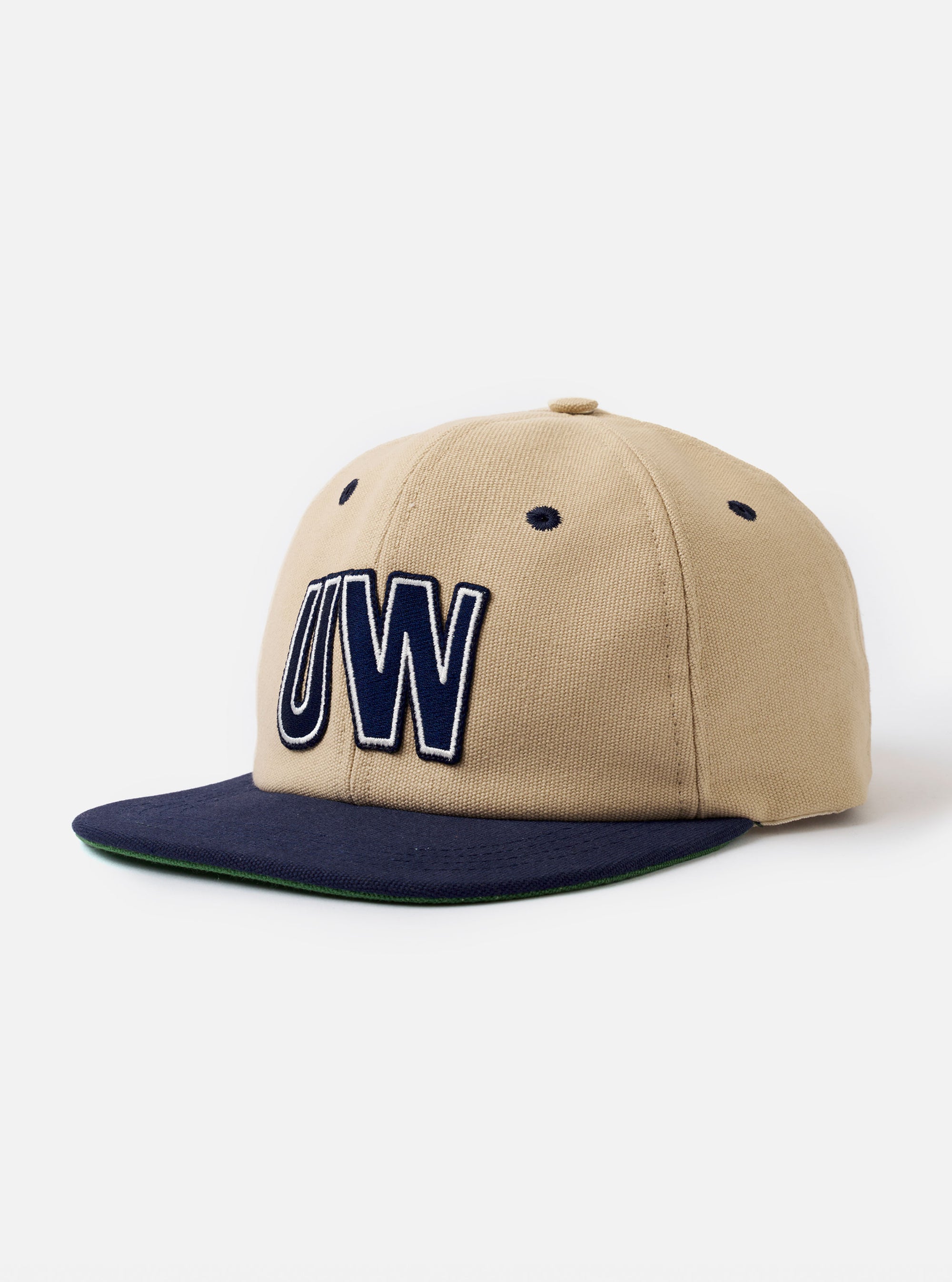 The Sand/Navy Canvas Baseball Hat is shown against a plain white background. The cap features a six-panel construction with a sand-colored crown and a contrasting navy blue curved brim. The design includes embroidered ventilation eyelets and a top button, all crafted from a durable canvas fabric.