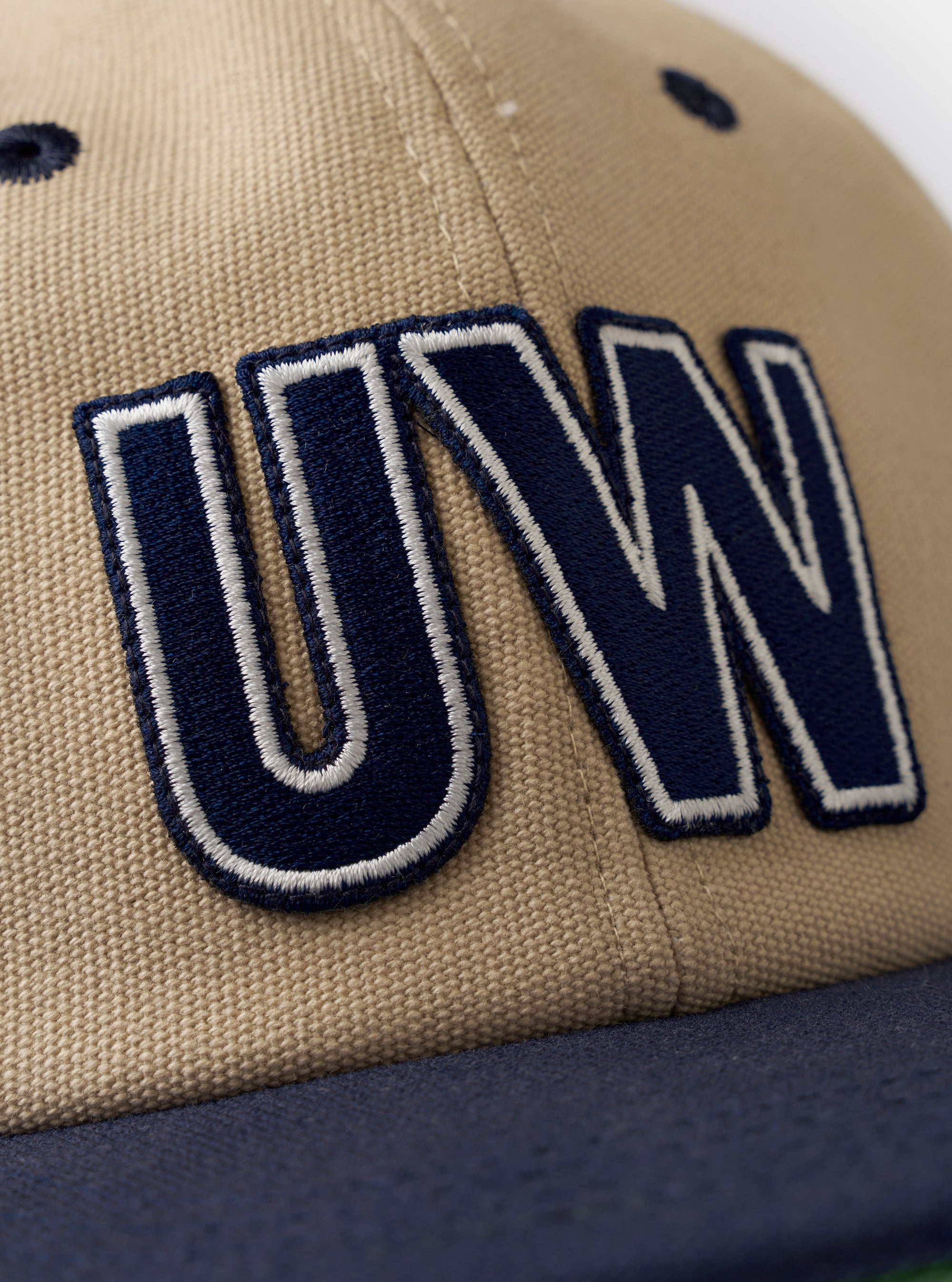 The Sand/Navy Canvas Baseball Hat is shown as a flat lay against a plain white background. The hat features a sand-colored canvas crown contrasted with a navy blue brim and a matching navy top button. It is constructed with six panels, tonal embroidered eyelets, and a curved peak.