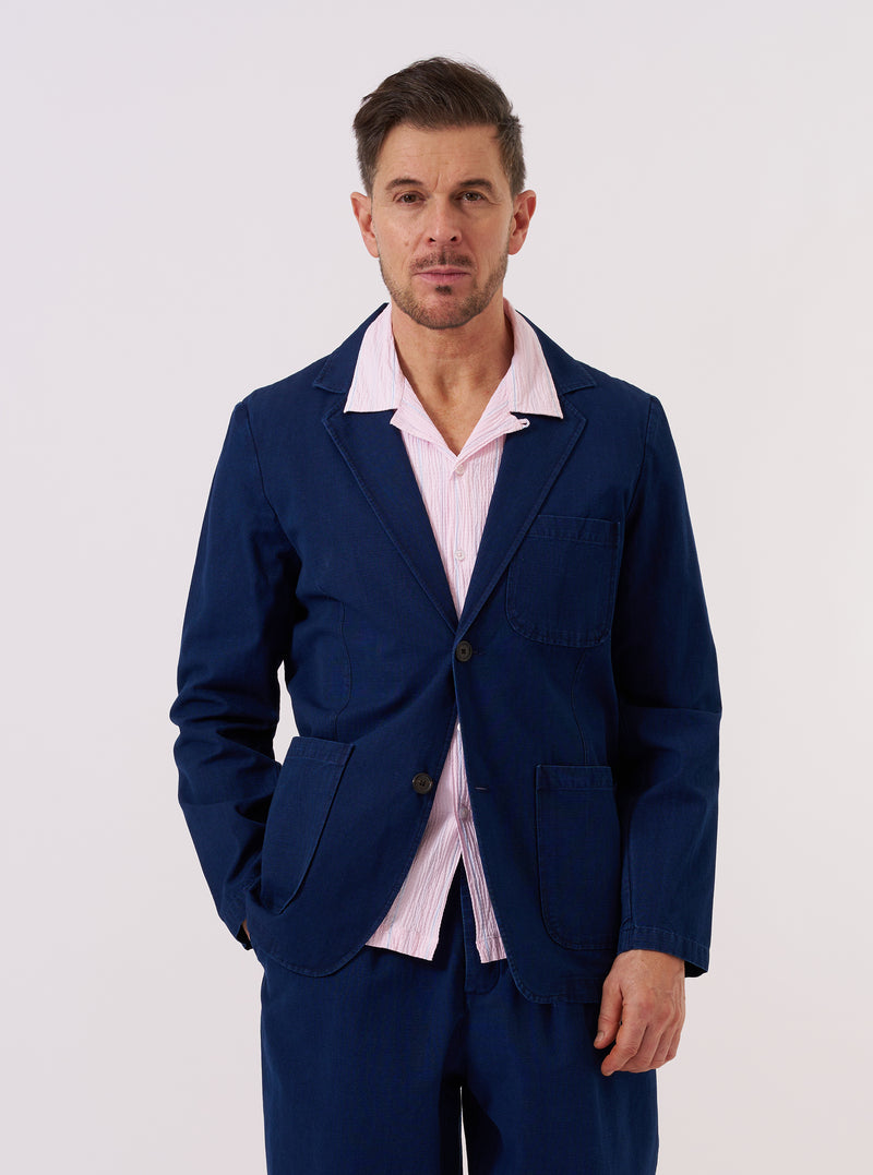 A model wears the Indigo Kimstop Two Button Jacket over a light-colored top. The jacket is a deep indigo blue with a relaxed fit, featuring a long, wide lapel, two-button front closure, a single chest patch pocket, and two large lower patch pockets. The image is a full-body shot taken outdoors against a blurred architectural background.