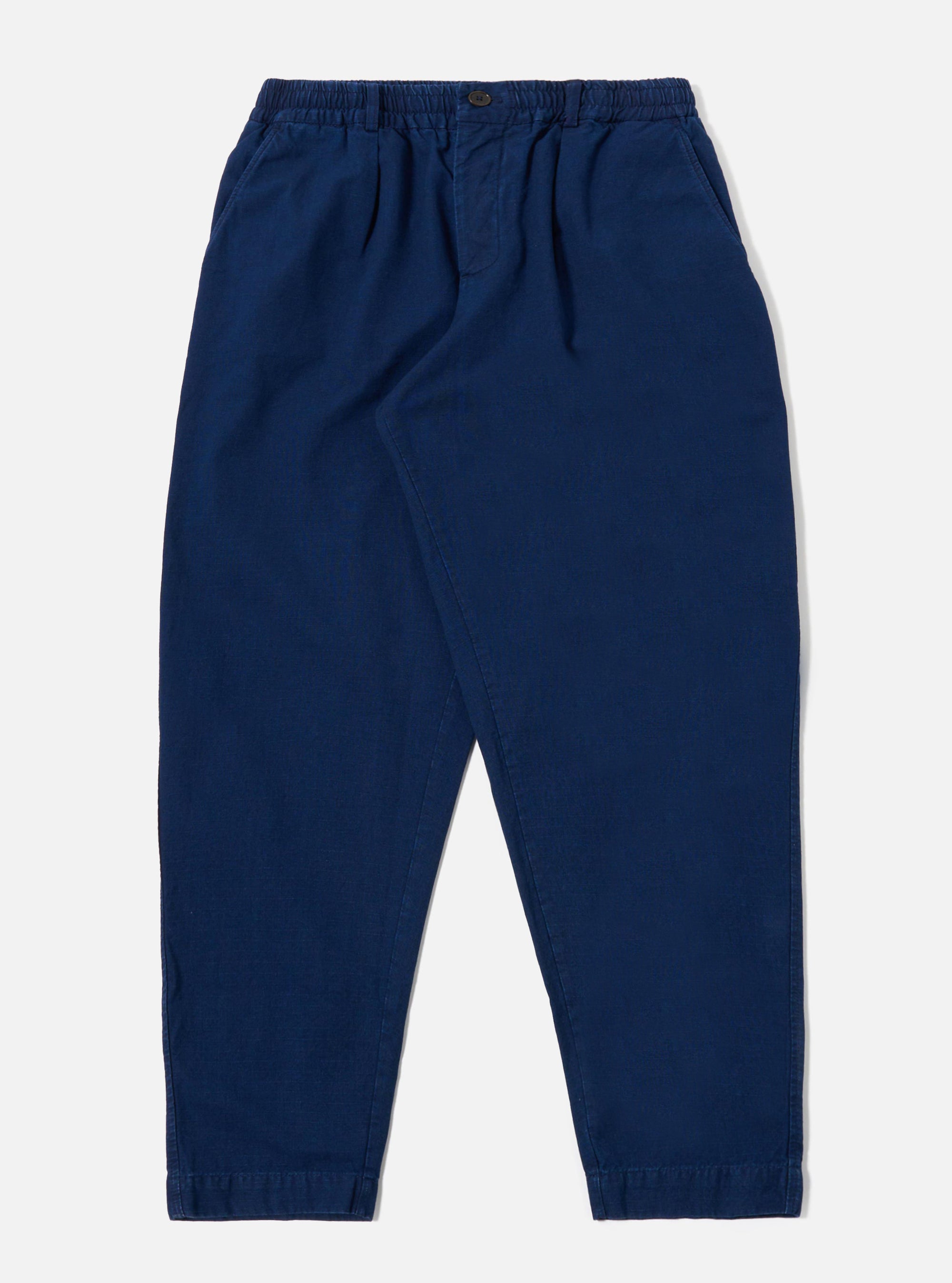 The Indigo Kimstop Pleated Track Pant is shown as a flat lay against a white background. The trousers feature an elasticated waistband with a drawstring, a single pleat at the front, and two side pockets. The fabric is a dark indigo organic cotton with a visible ripstop texture.