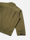 A model wears the Walsh x Universal Works Light Olive Twill Troopers Jacket shown from a three-quarter angle against a light background. The photo emphasizes the large front bellows pockets with buttoned flaps and adjustable cuffs.
