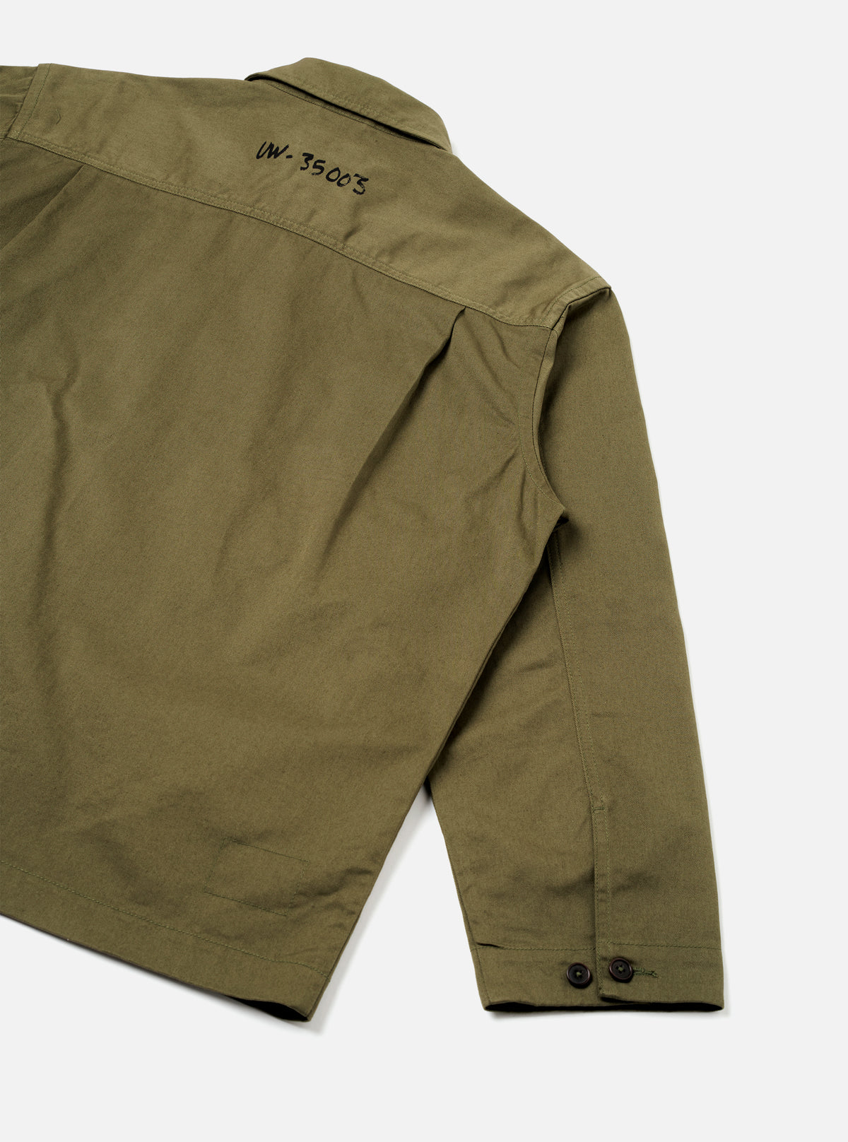 A model wears the Walsh x Universal Works Light Olive Twill Troopers Jacket shown from a three-quarter angle against a light background. The photo emphasizes the large front bellows pockets with buttoned flaps and adjustable cuffs.