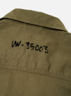 A close-up view of the Walsh x Universal Works Light Olive Twill Troopers Jacket worn by a model against a studio backdrop. The shot focuses on the durable twill weave, contrast stitching around pockets, collar structure, and buttons.