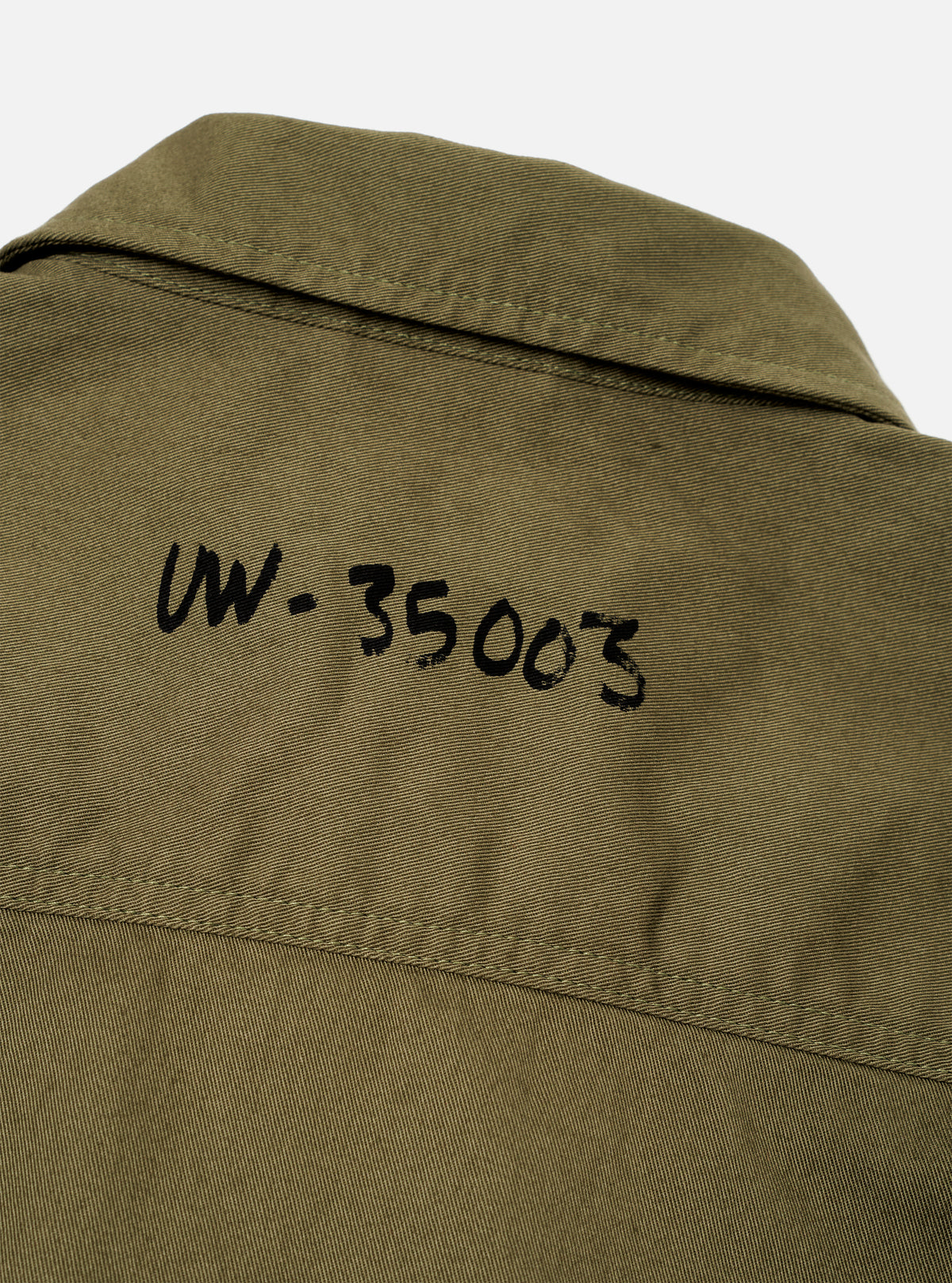 A close-up view of the Walsh x Universal Works Light Olive Twill Troopers Jacket worn by a model against a studio backdrop. The shot focuses on the durable twill weave, contrast stitching around pockets, collar structure, and buttons.