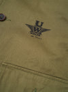Detailed view of the Walsh x Universal Works Light Olive Twill Troopers Jacket in a studio setting. The photo highlights the fine cotton twill texture, chest seam details, button placket, and overall workwear-inspired craftsmanship.