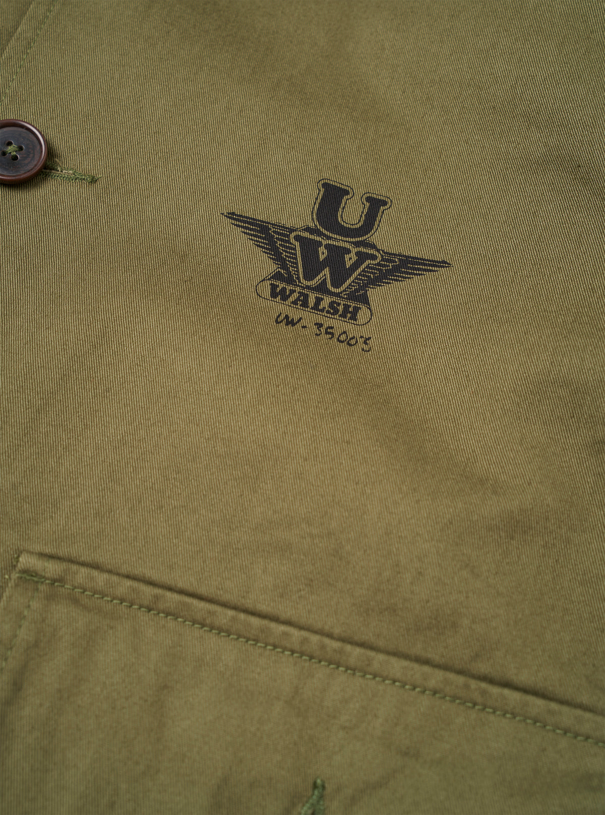 Detailed view of the Walsh x Universal Works Light Olive Twill Troopers Jacket in a studio setting. The photo highlights the fine cotton twill texture, chest seam details, button placket, and overall workwear-inspired craftsmanship.