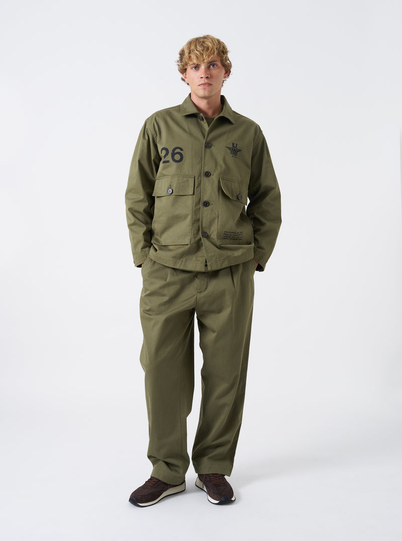 A model wears the Walsh x Universal Works Light Olive Twill Troopers Jacket against a plain neutral background. The full shot highlights the relaxed fit, utility button pockets, collared neckline, and light olive twill fabric.