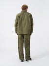 A model stands wearing the Walsh x Universal Works Light Olive Twill Troopers Jacket against a plain studio background. The side perspective emphasizes the structured fit, sleeve details, buttoned cuffs, and light olive green color.