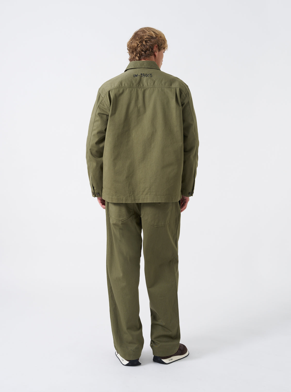 A model stands wearing the Walsh x Universal Works Light Olive Twill Troopers Jacket against a plain studio background. The side perspective emphasizes the structured fit, sleeve details, buttoned cuffs, and light olive green color.