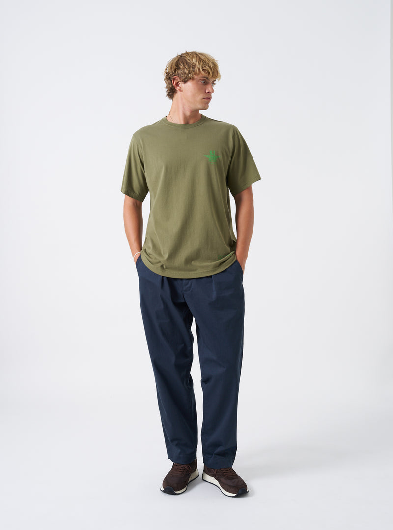 A model wears the Walsh x Universal Works Olive Single Jersey T Shirt in front of a plain background. The shot highlights the comfortable crewneck cut, short sleeve design, smooth olive green cotton jersey texture, and casual styling.
