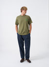 A model wears the Walsh x Universal Works Olive Single Jersey T Shirt in front of a plain background. The shot highlights the comfortable crewneck cut, short sleeve design, smooth olive green cotton jersey texture, and casual styling.