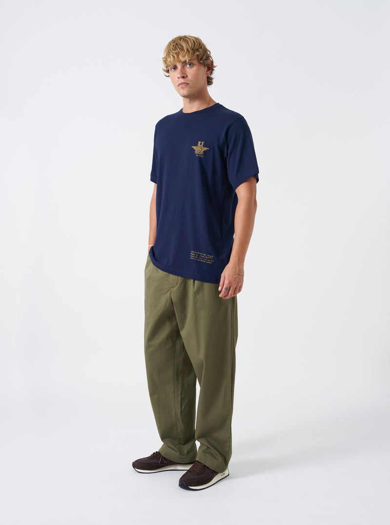 A model stands against a neutral backdrop wearing the Walsh x Universal Works Navy Single Jersey T Shirt. The shot displays the short sleeves, crew neckline, soft jersey fabric, dark navy blue tone, and casual silhouette.