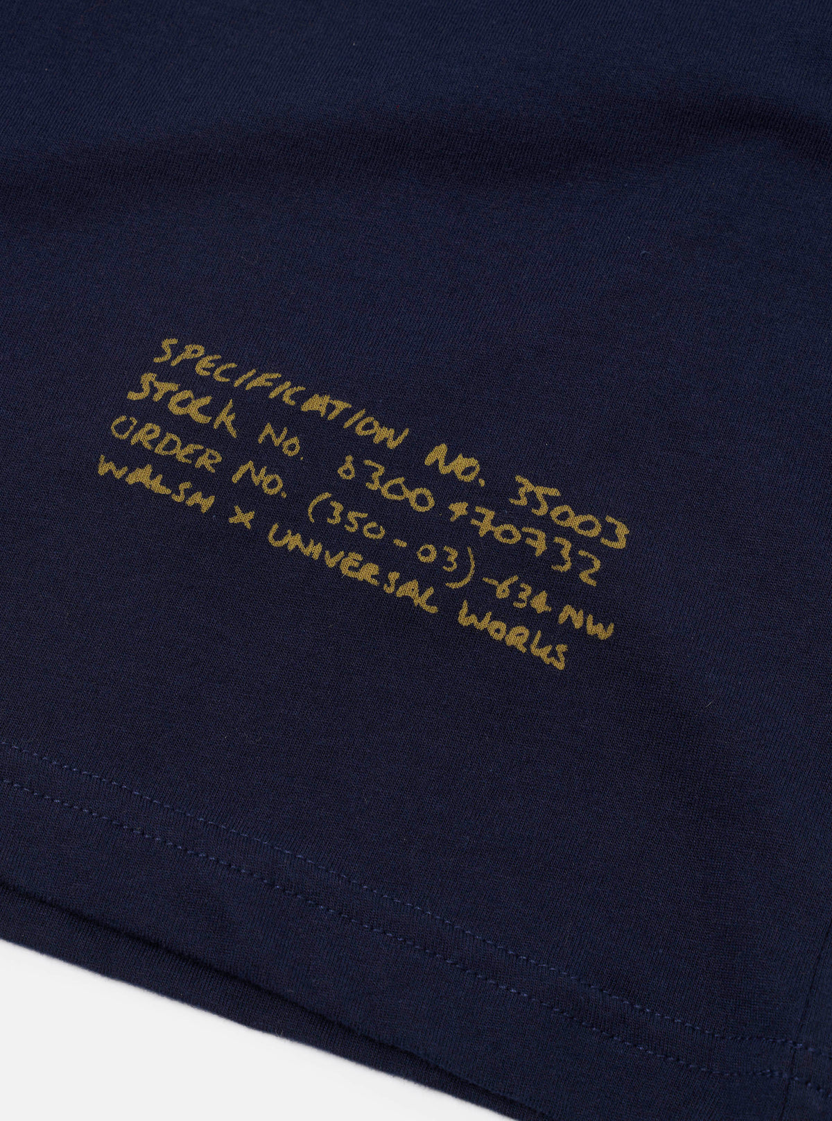 A close-up view of the collar and print detail on the Walsh x Universal Works Navy Single Jersey T Shirt against a neutral background. The image emphasizes the fine ribbing, collar construction, logo graphics, and navy cotton fabric.