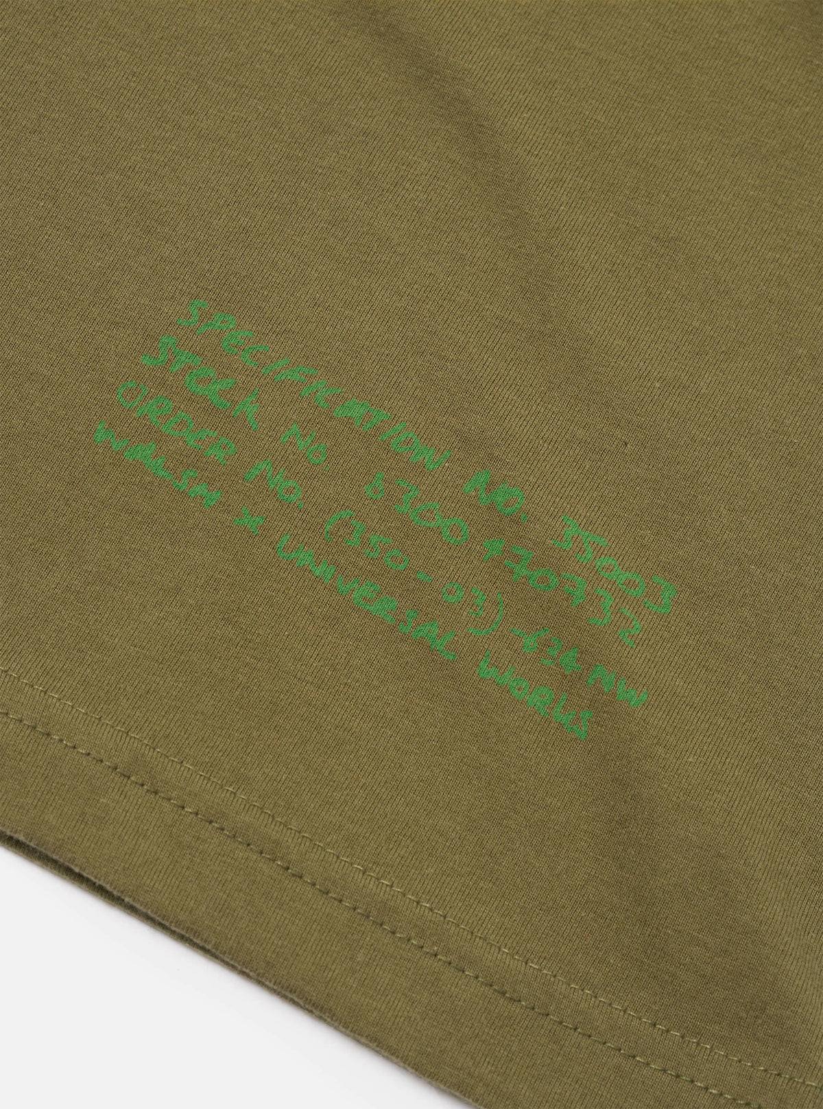 A close-up shot focusing on the collar and chest graphics of the Walsh x Universal Works Olive Single Jersey T Shirt against a plain background. The image showcases the detailed stitching, graphic print, and olive jersey fabric.